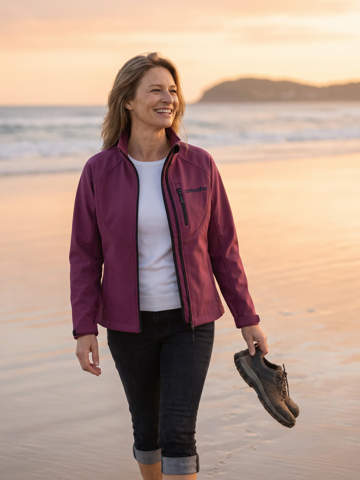 A woman walking a wide beach at sunset with her shoes in her hand, wearing the berry Softshell